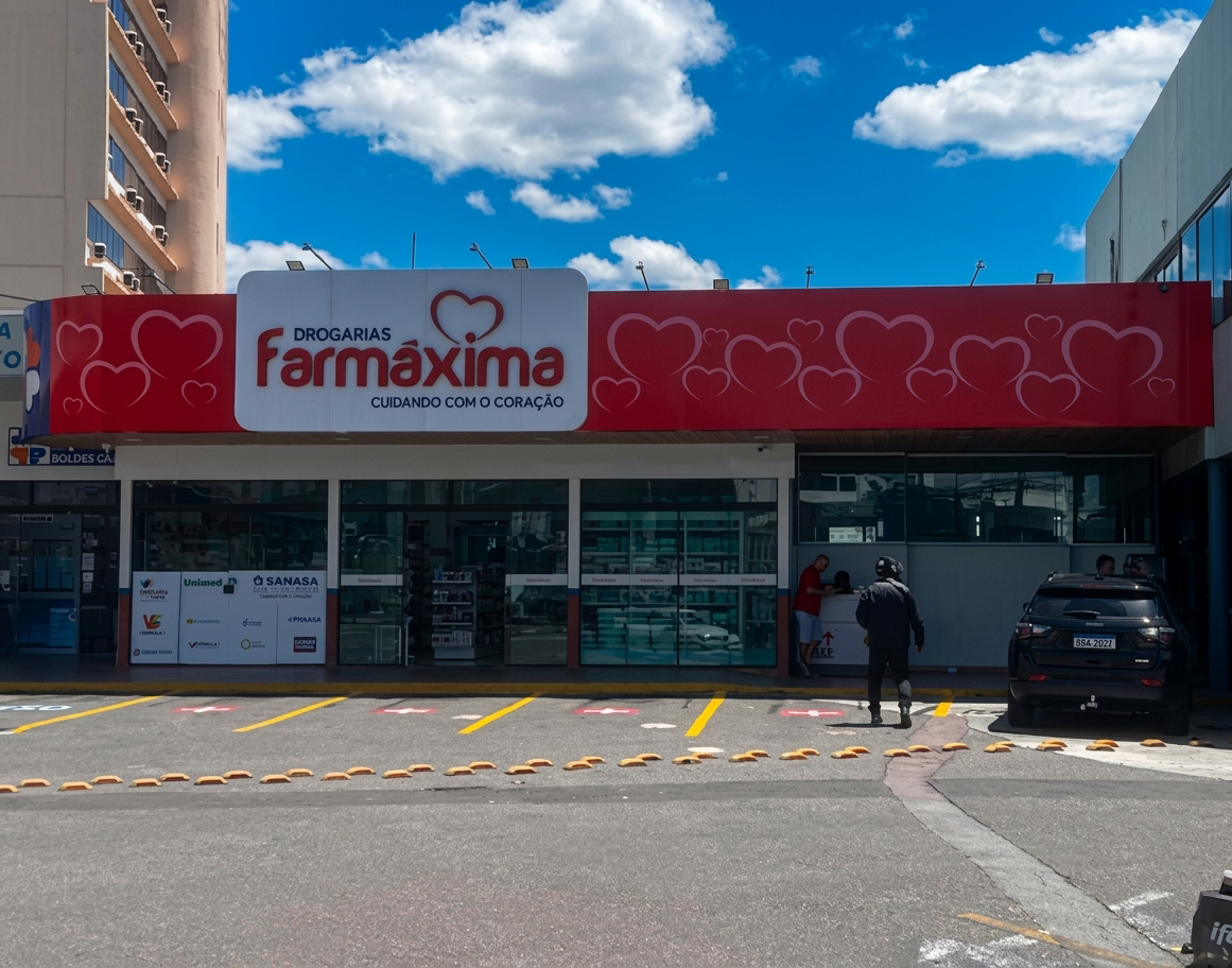 Vista lateral da loja Farmáxima, com prateleiras e produtos visíveis pela vitrine